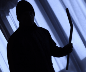 A shadowy figure in a beanie holding a crowbar, standing in front of a curtained window.