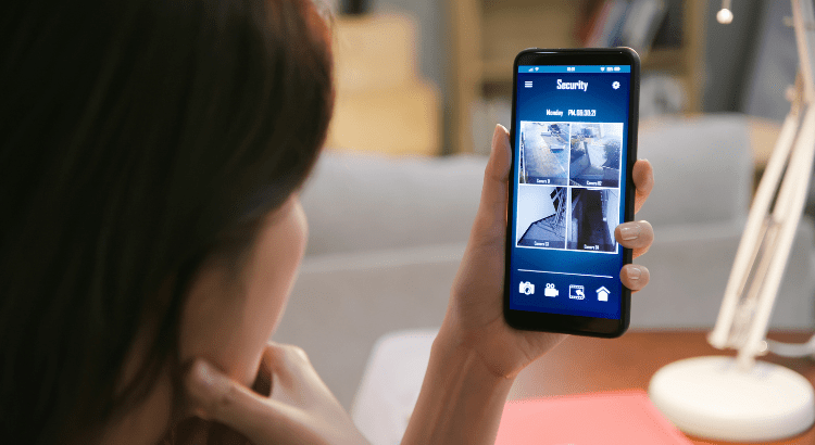 A woman holding a smartphone, viewing the live feed from multiple home security cameras on the screen.