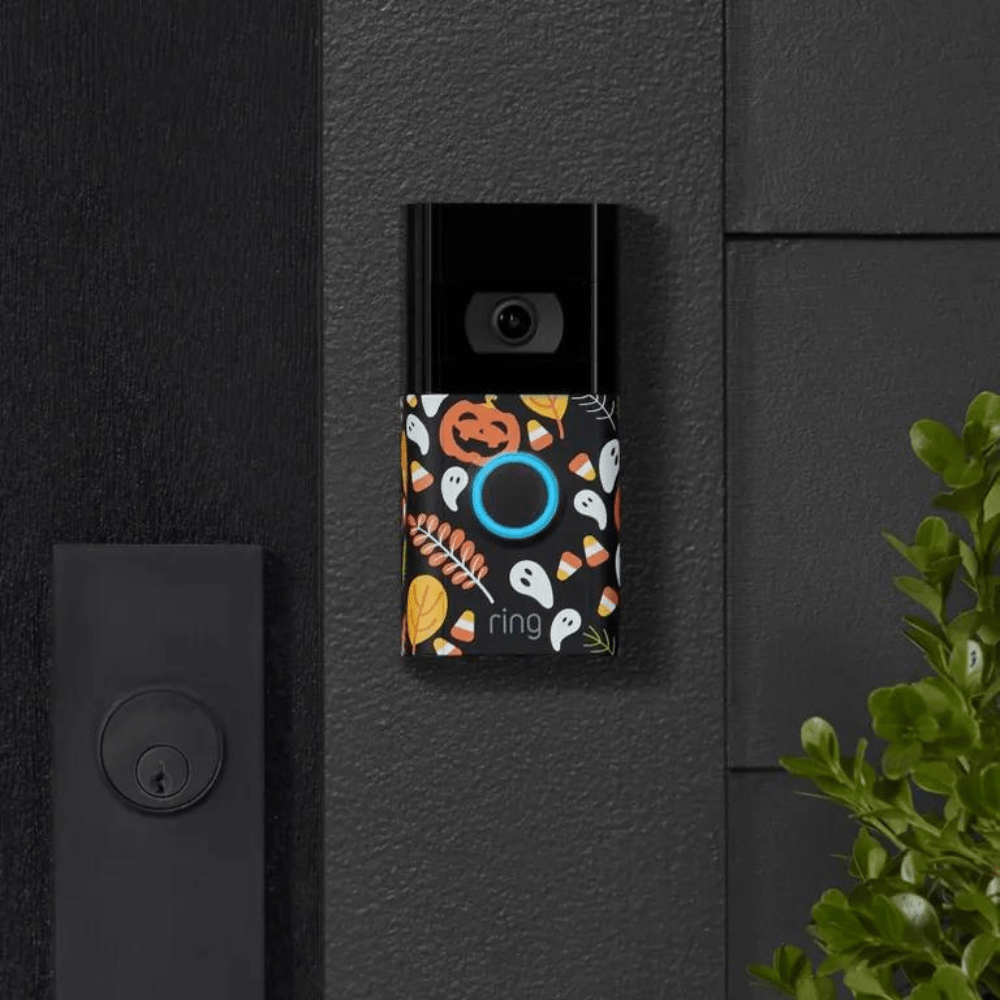 Ring Doorbell camera adorned with a festive Halloween faceplate featuring pumpkins, candy corn, ghosts, and fall leaves. The doorbell is mounted on a dark wall next to a door handle, with greenery partially visible on the right side.