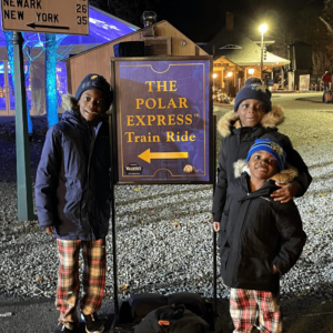 Three children smiling at the camera, standing next to a sign for "The Polar Express Train Ride" during a nighttime event, dressed warmly in winter clothing.