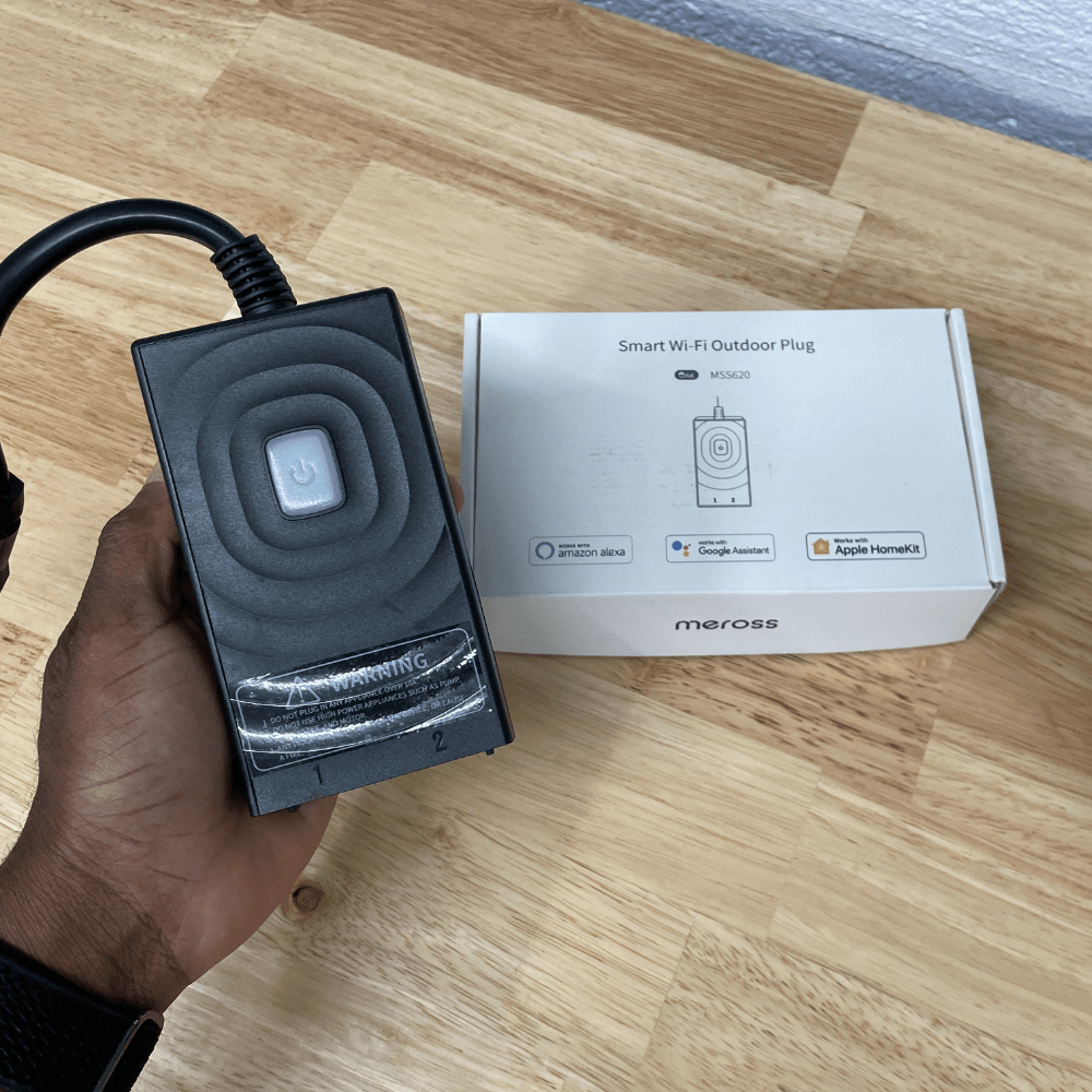 A hand holding a Meross Smart Wi-Fi Outdoor Plug, with its box in the background. The plug is designed for outdoor use and compatible with Amazon Alexa, Google Assistant, and Apple HomeKit.